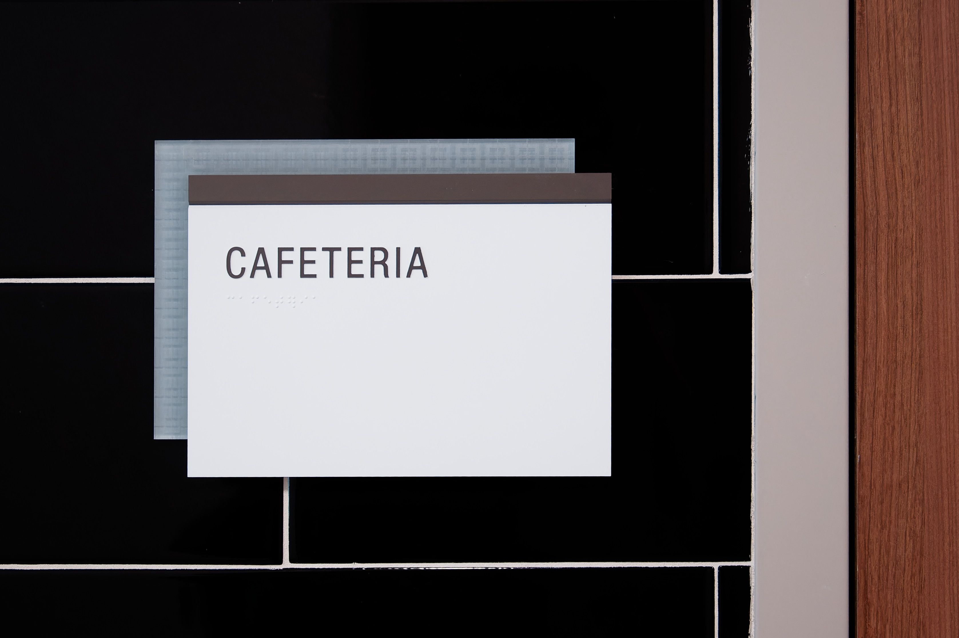 A room sign with tactile "CAFETERIA" text and braille, with a laser etched patterned acrylic accent panel, is mounted on a tiled wall.