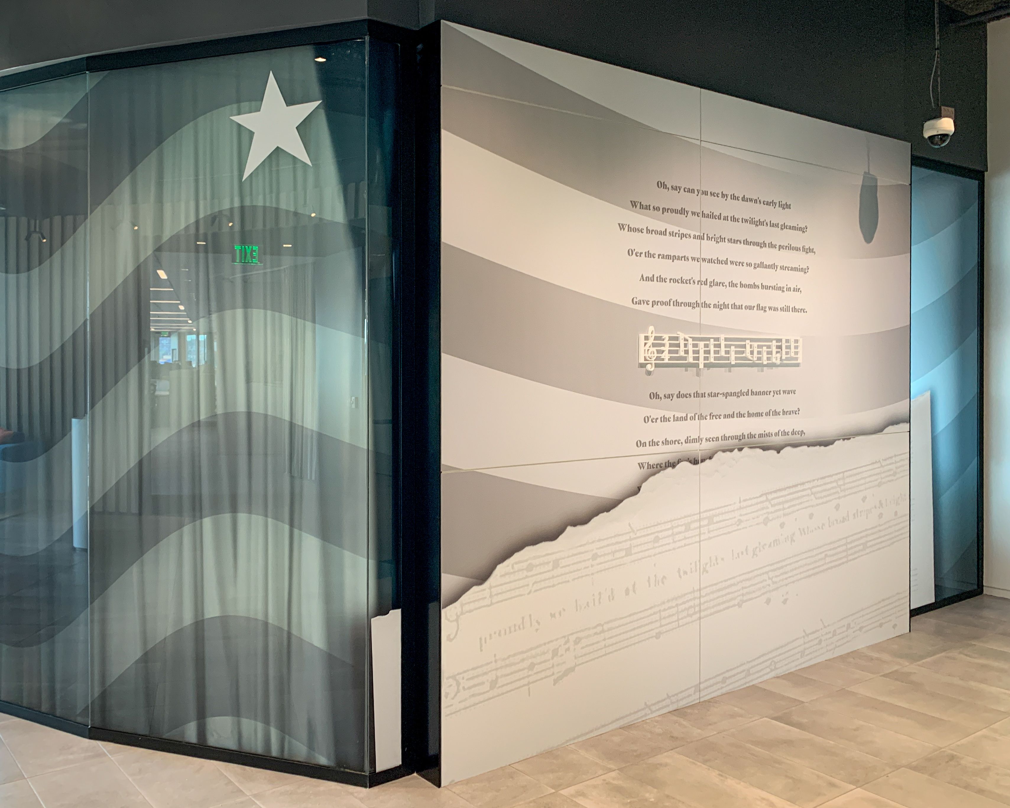 A graphic of flag stripes and stars is applied across several glass panels and a wall in a corporate office. On the wall is a printed graphic of sheet music and words to the Star Spangled Banner, and an interactive musical xylophone where users can play the first line of the song.