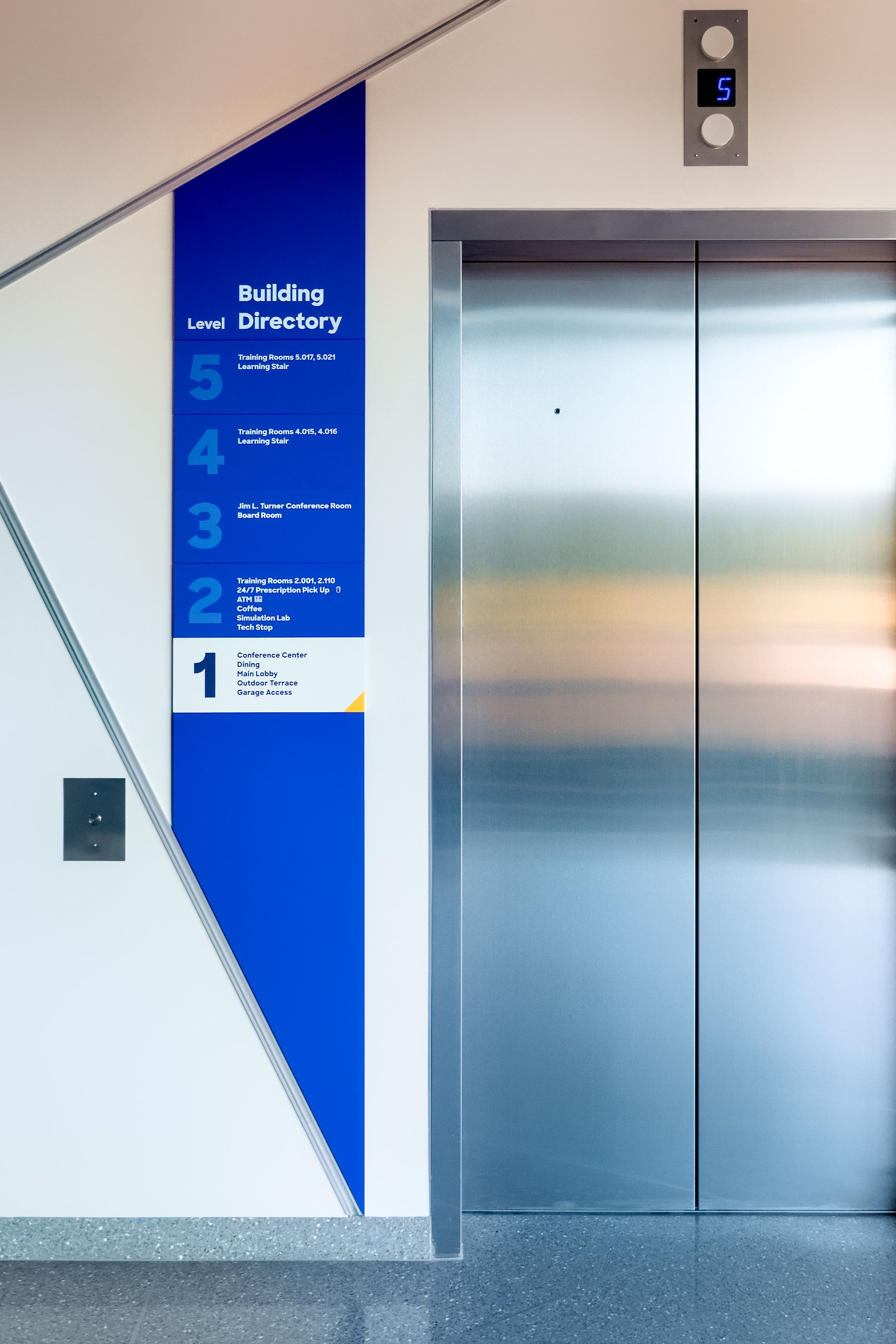 A large blue building directory sign mounted on a wall next to an elevator shows the names of various departments and their levels. The sign has angular top and bottom edges mtaching aluminum reveals cut into the wall.