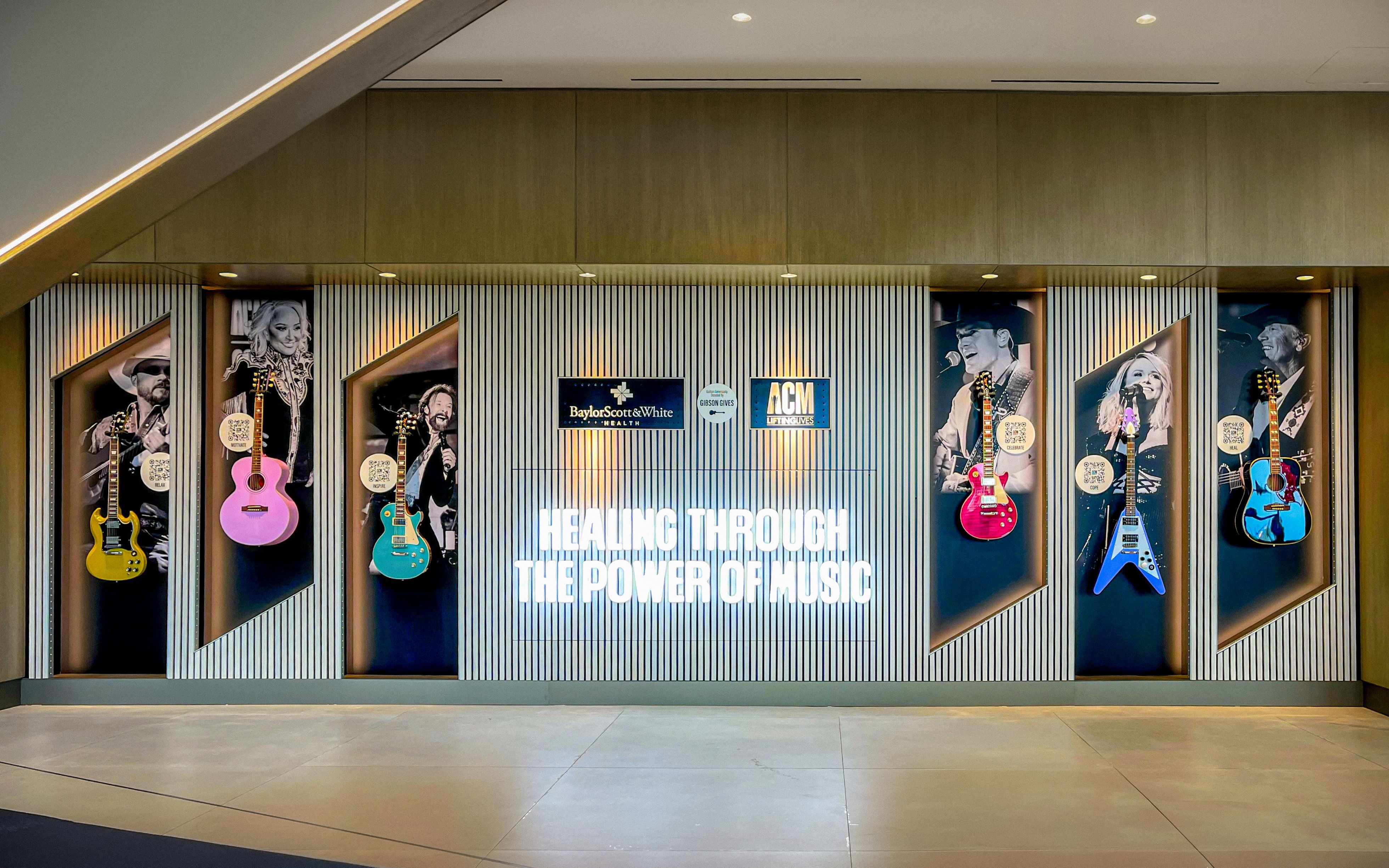 A feature wall in a hopsital with glowing LED letters saying "HEALING THROUGH THE POWER OF MUSIC". Inset into the wall are large printed graphics of various country music stars, with real guitars displayed and QR codes inviting the users to learn more.