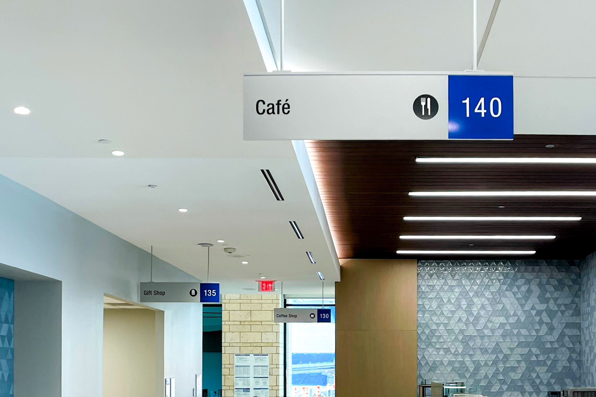 Silver signs for a Café, Gift Shop, and Coffee Shop hang from the ceiling in a hospital. They have a blue accent and vinyl cut symbols.