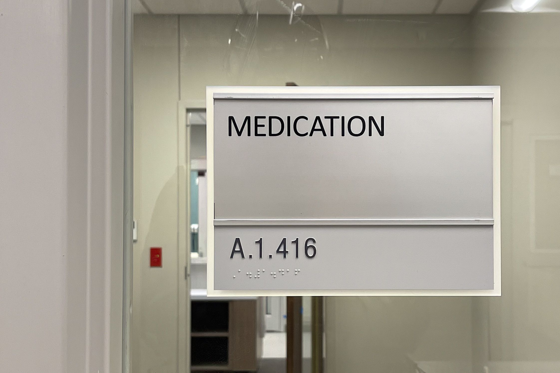 A silver and white medication room sign mounted on glass in a hospital, displaying the room number in raised text and braille.