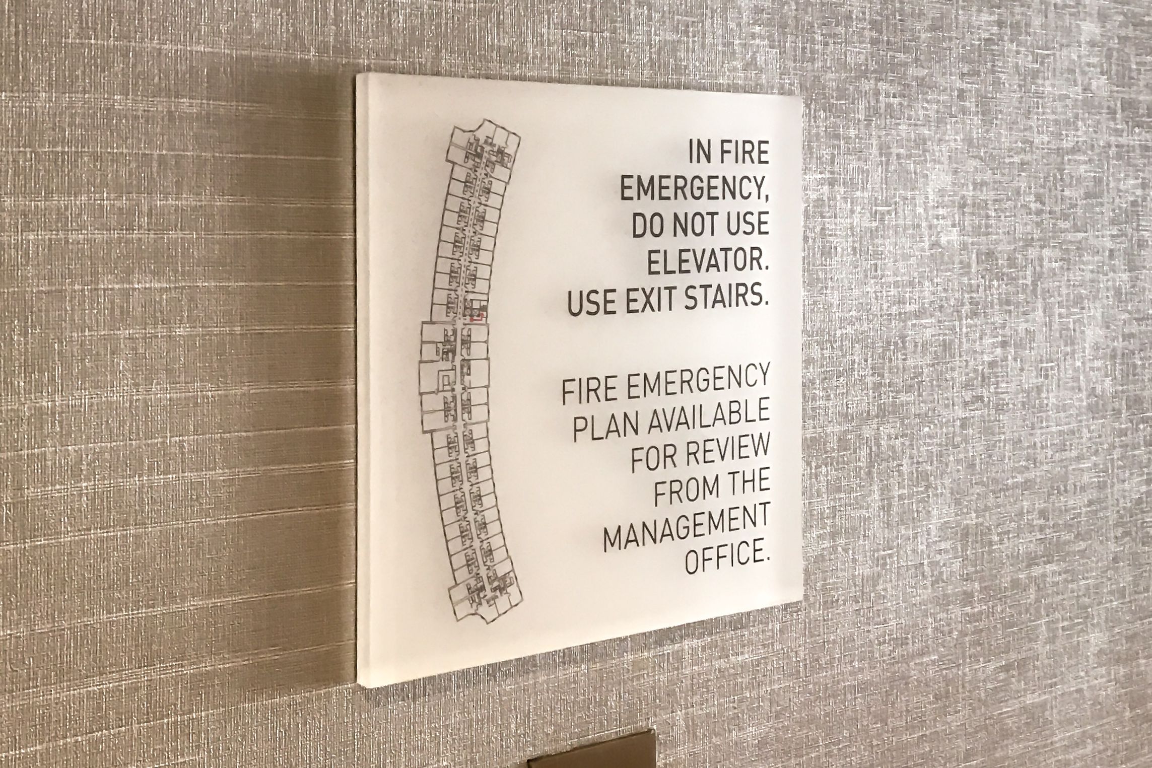 A frosted acrylic sign with a direct UV printed evacuation plan mounted on the wall above an elevator call button.