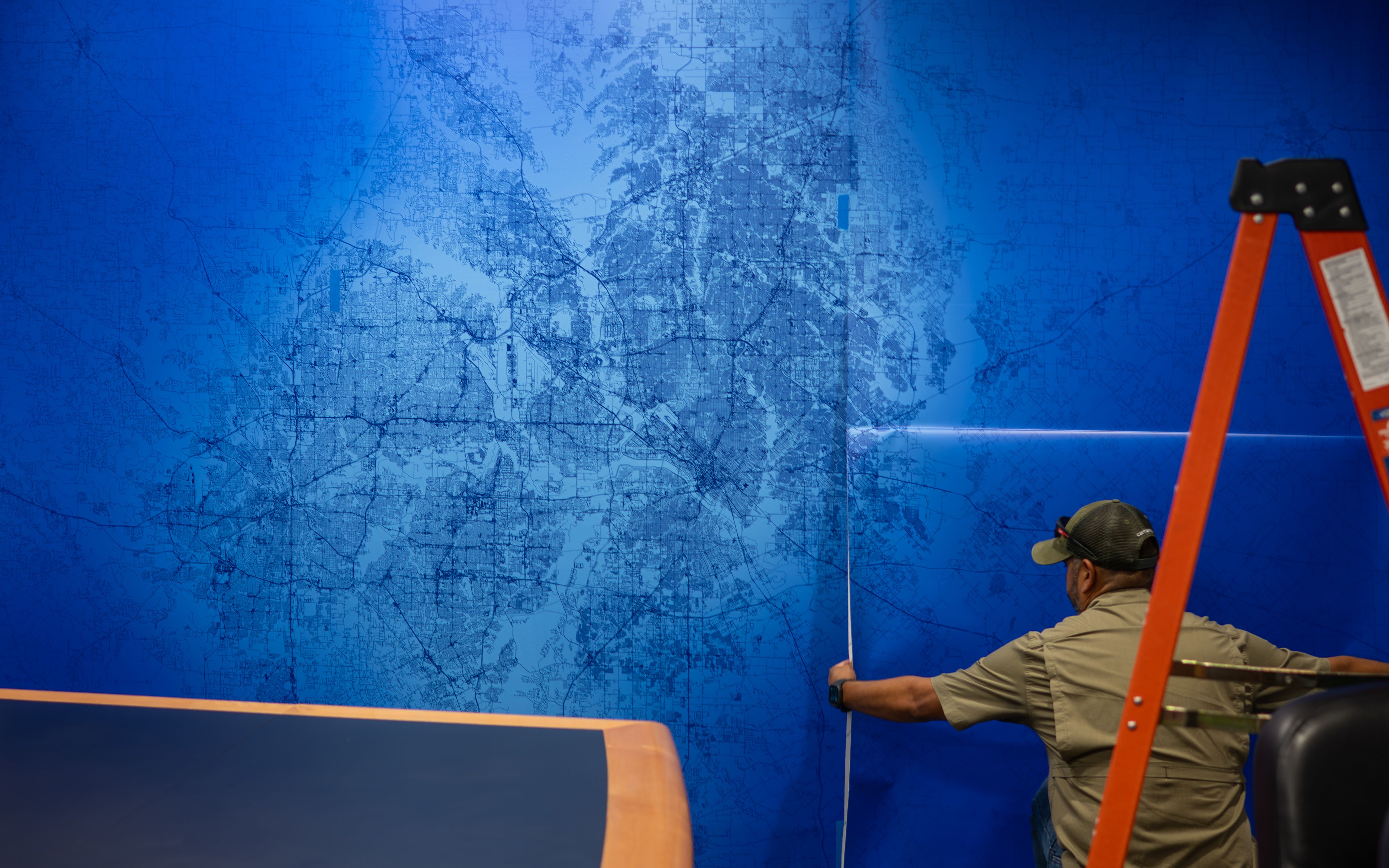 A graphic installer is applying a large vinyl graphic map of the Dallas-Fort Worth Metroplex to a wall in a conference room.
