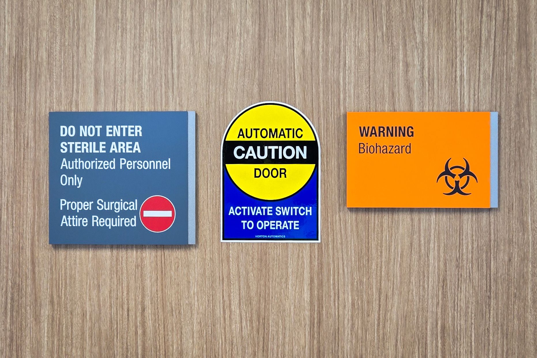 A pair of signs are mounted on a door in a hospital. One is grey and silver with text stating not to enter and alerting that a sterile area is beyond the door. The other is bright orange and silver with a biohazard symbol and warning text.