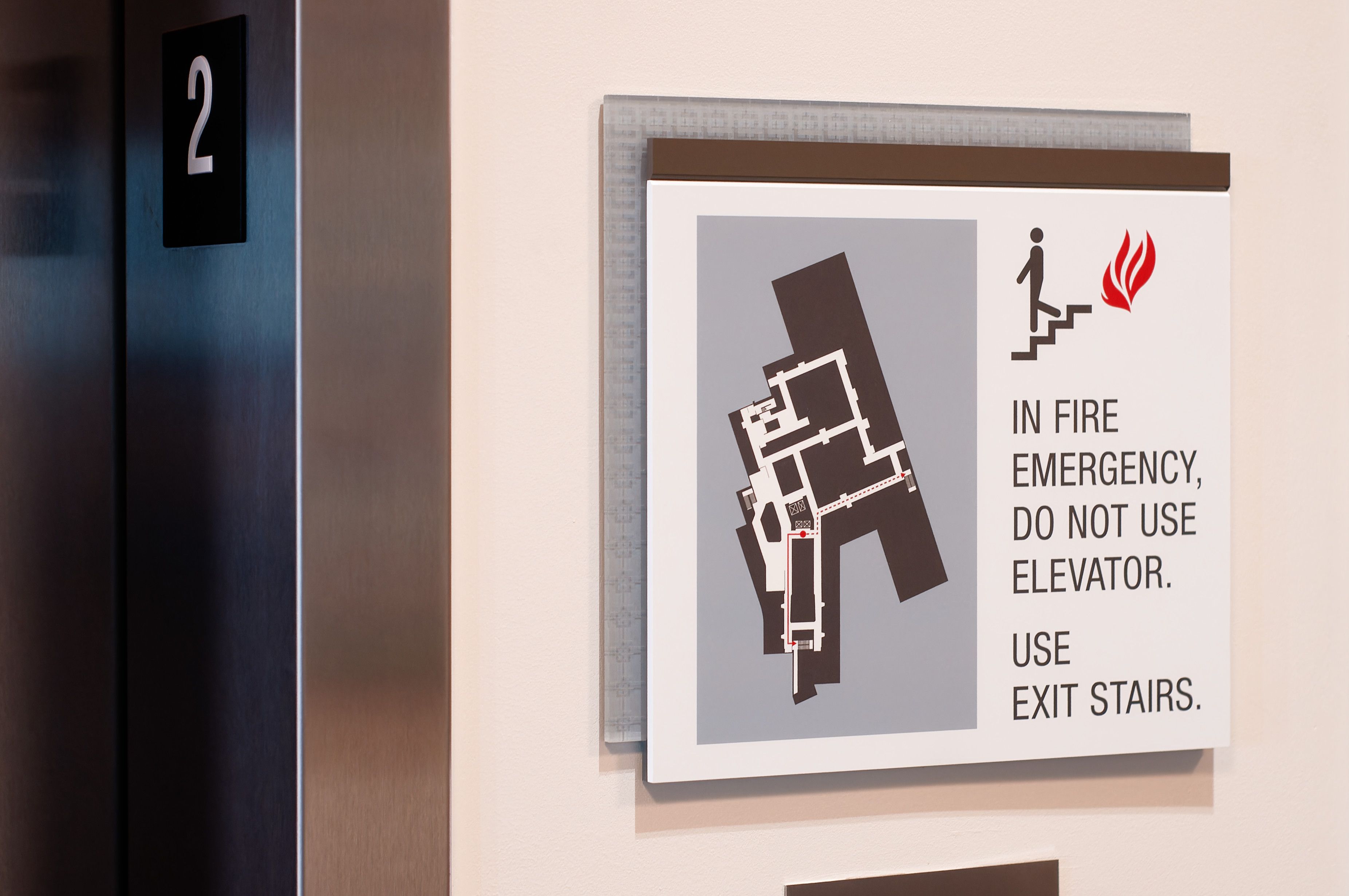 A sign mounted above an elevator call button in a hospital shows a printed fire evacuation plan and states to use the stairs in case of fire. It's accented with a light blue laser etched patterned acrylic panel and brown header.