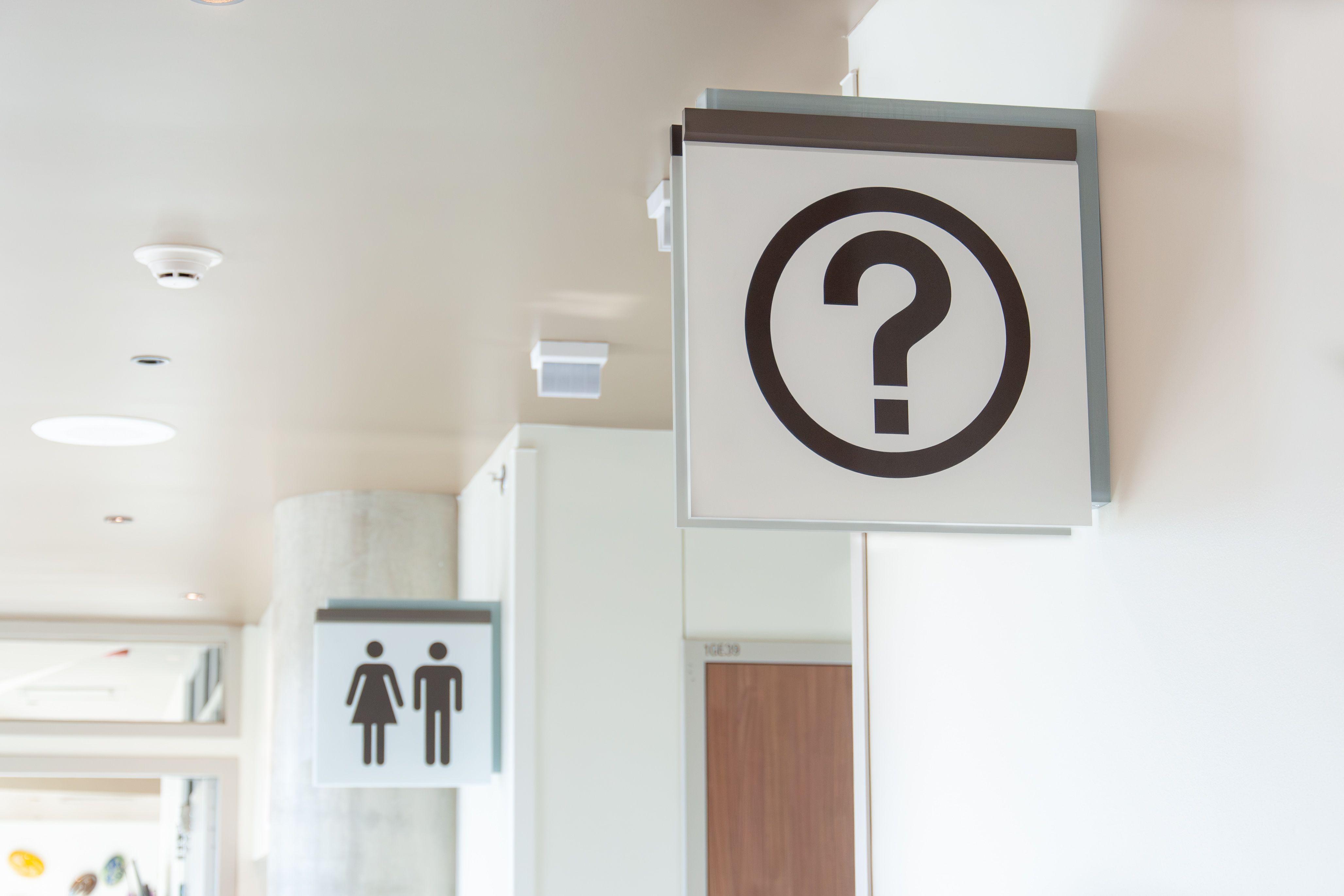 Two projecting flag signs are mounted on a wall in a hospital lobby. One has a brown vinyl restroom symbol and the other has a brown question mark symbol.
