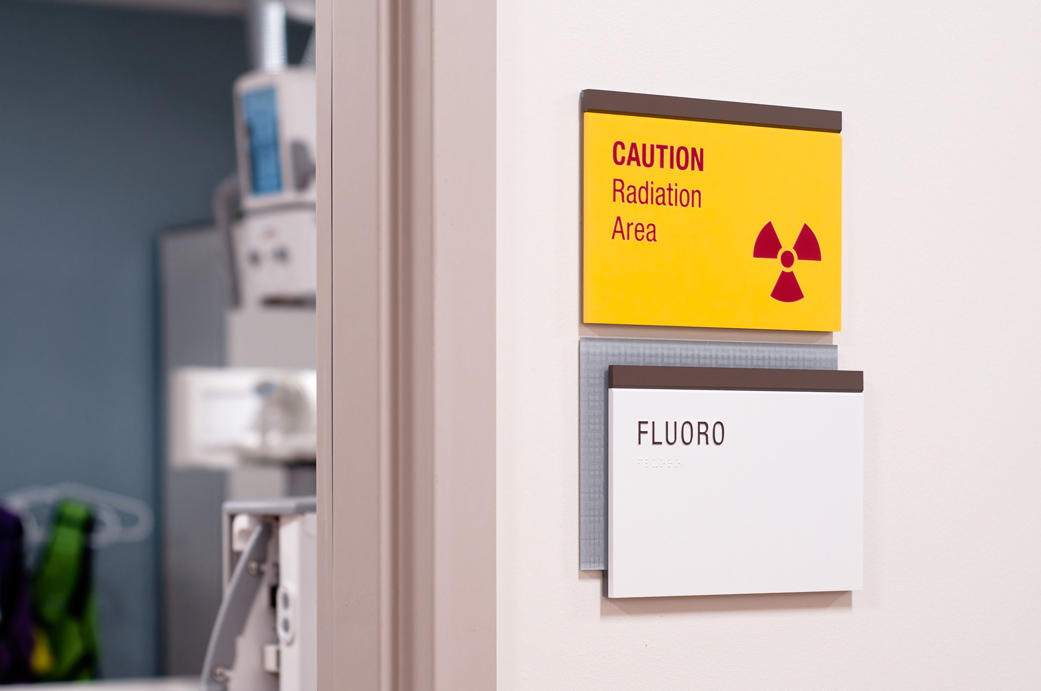 A tactile room sign with a bright yellow Caution Radiation notice sign above is located on the wall outside of a Fluoroscopy room.