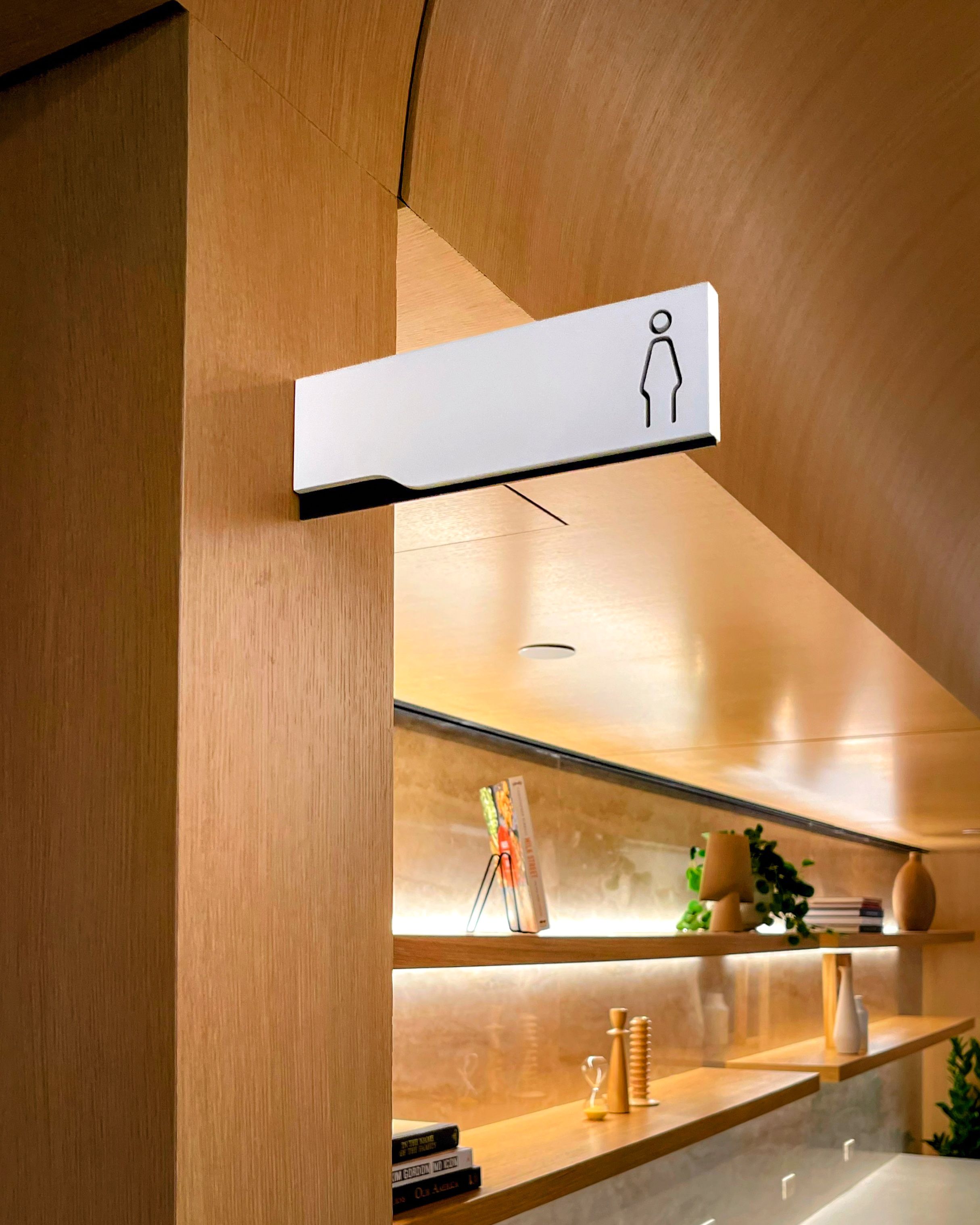 A white and black flag sign with a laser cut restroom symbol and simple, modern design projects from a wooden wall in an office building.