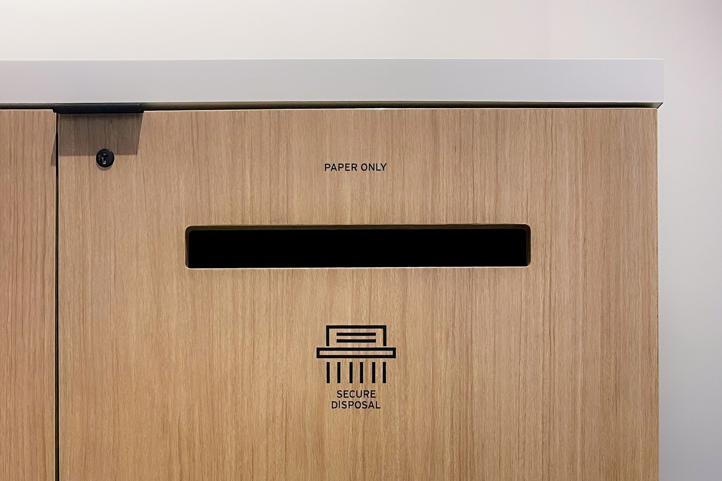 Cut black vinyl text stating "PAPER ONLY" and "SECURE DISPOSAL", and a modern symbol of paper shredding, is applied to a wooded cabinet door with a slot for paper disposal.