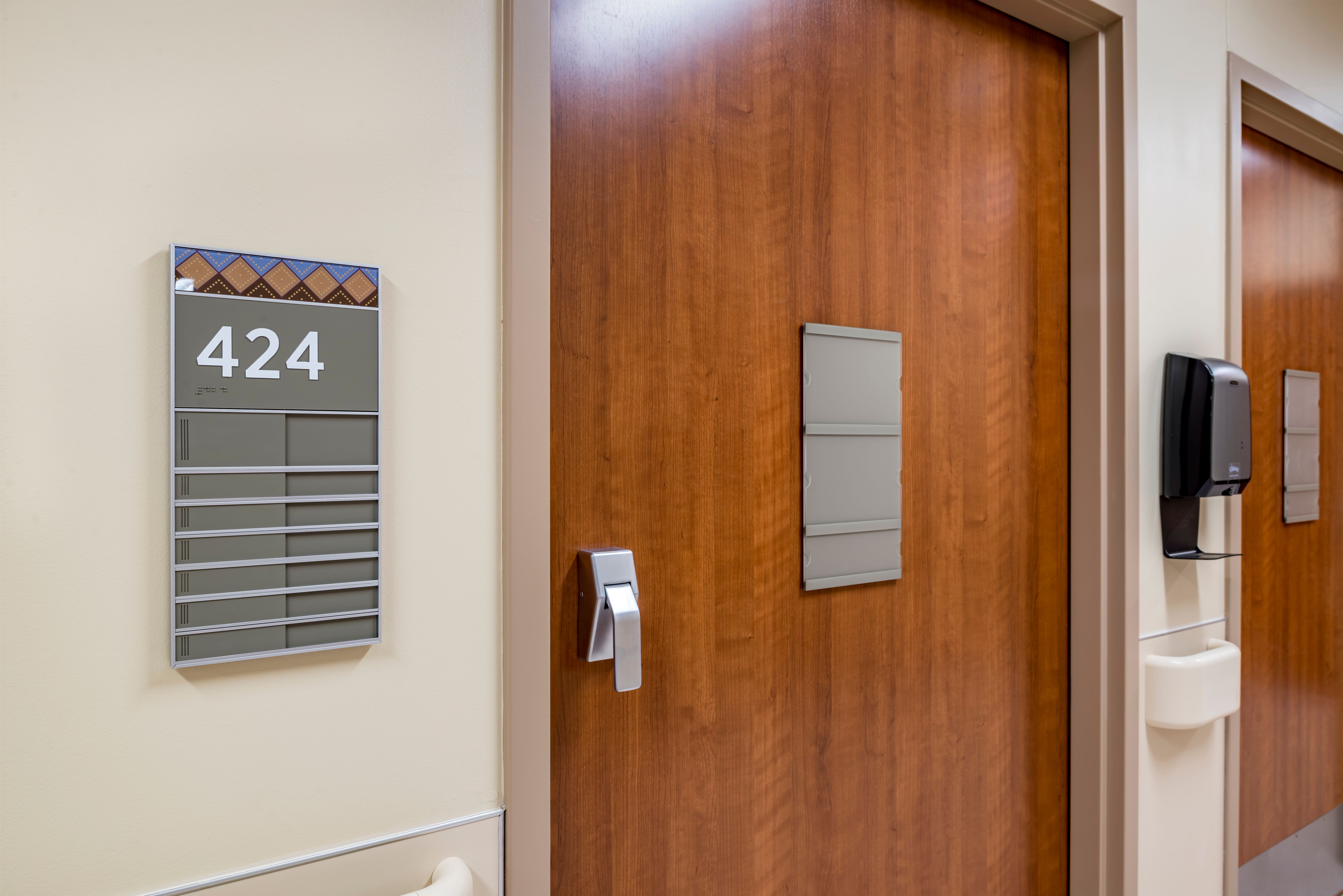 A patient room sign with a tactile room number, braille, and patient notice sliders, next to a hospital room door