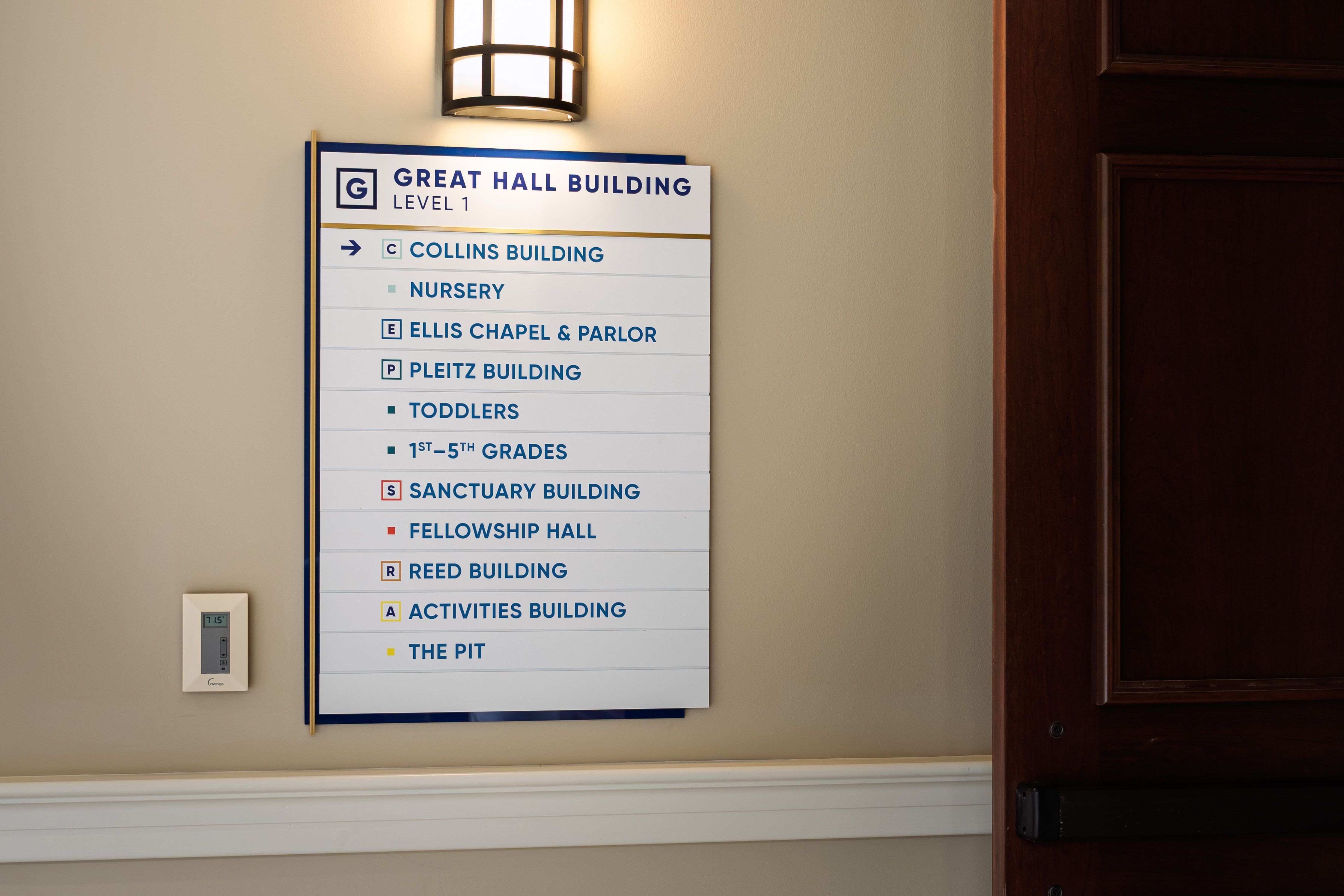 A wall-mounted directional sign with a brushed brass accent bar and blue background panel directs to the various destinations in a church building. At the top is a header that states "Great Hall Building Level 1"