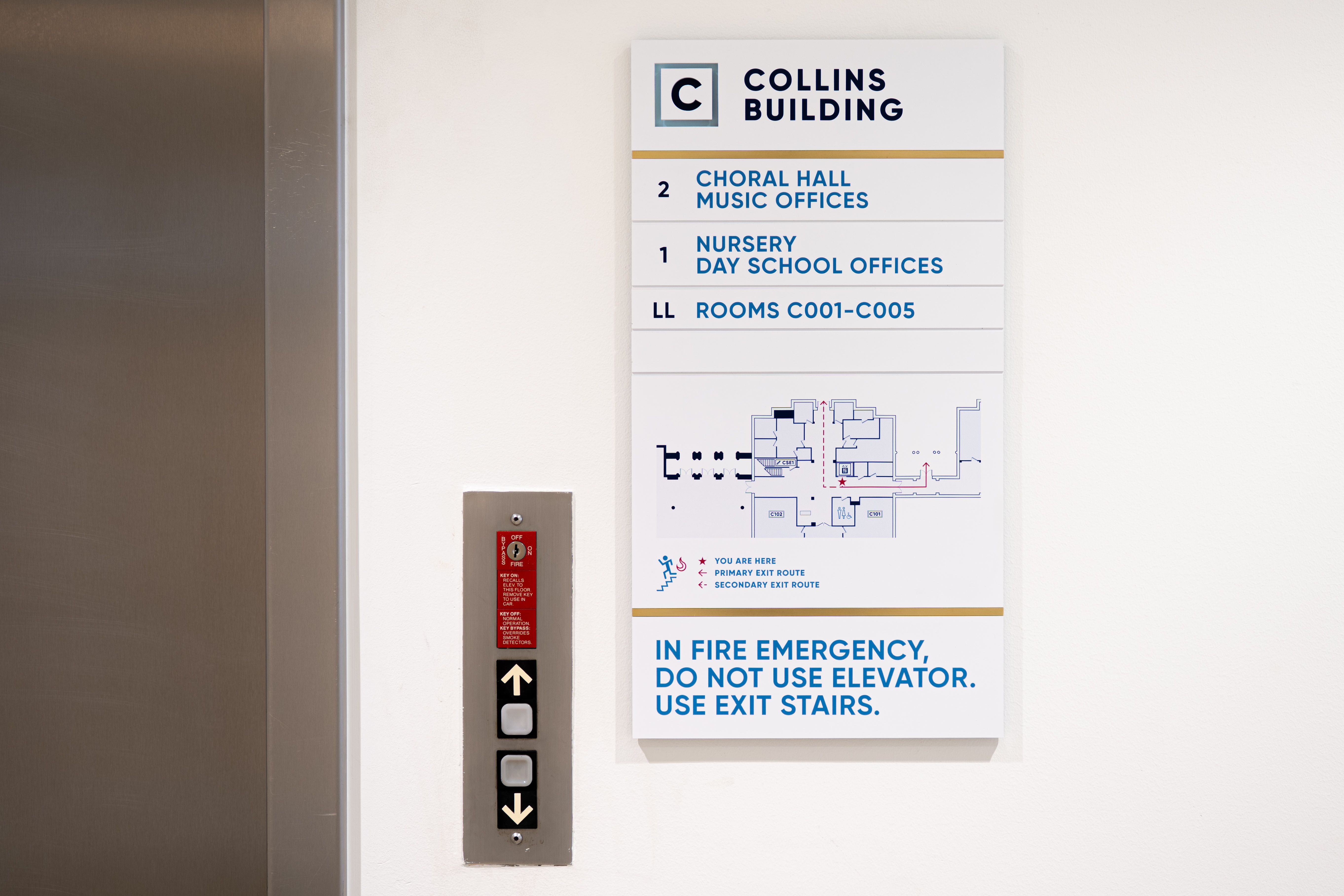 A combined elevator evacuation map sign and directory listing level destinations are mounted next to an elevator call button. The sign has brushed brass accents and indicates the viewer is in the "Collins Building"