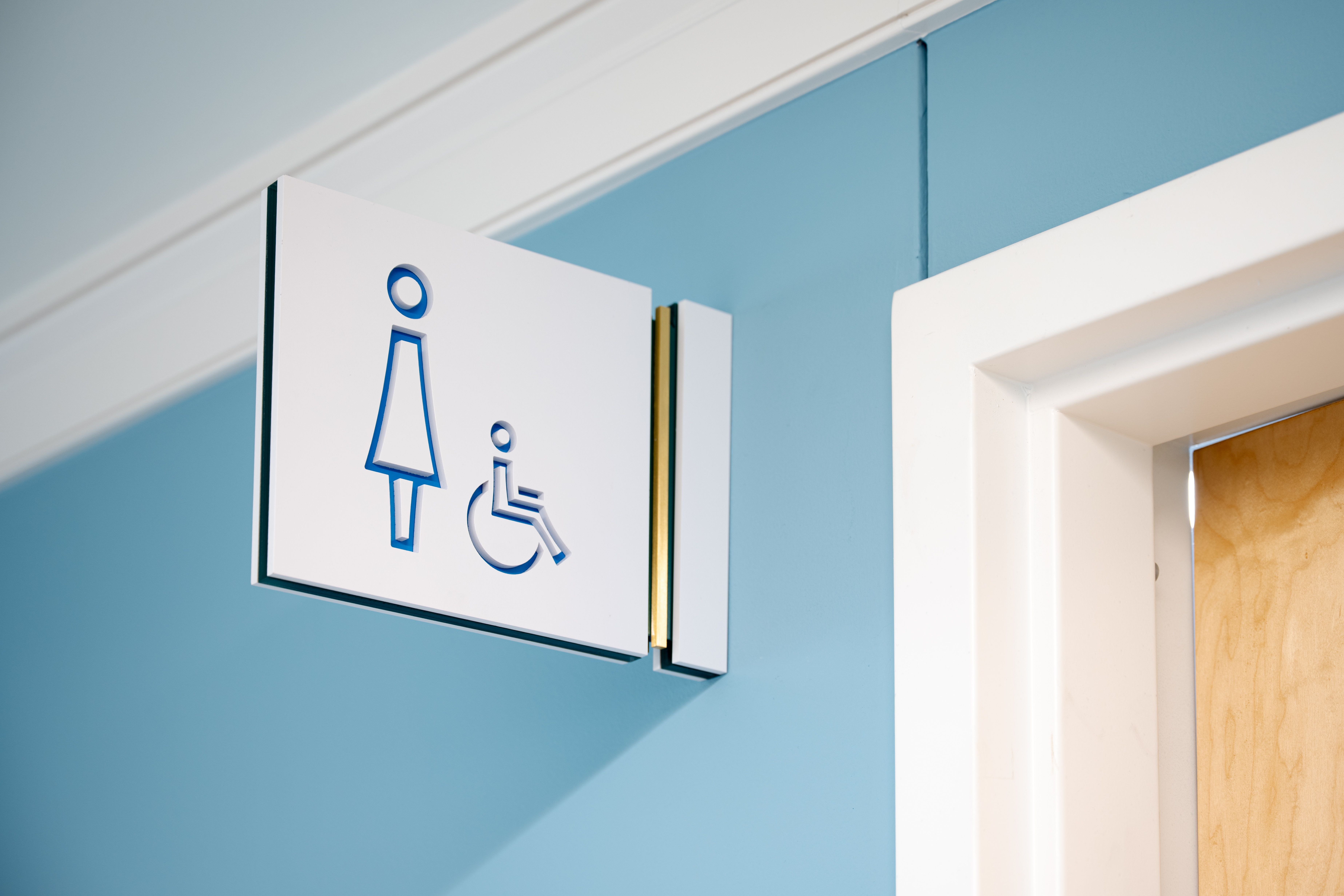 A white projecting flag sign with a women's restroom symbol cut out of it is mounted on a blue wall. The sign has a green accent inner layer and a brushed brass accent strip.