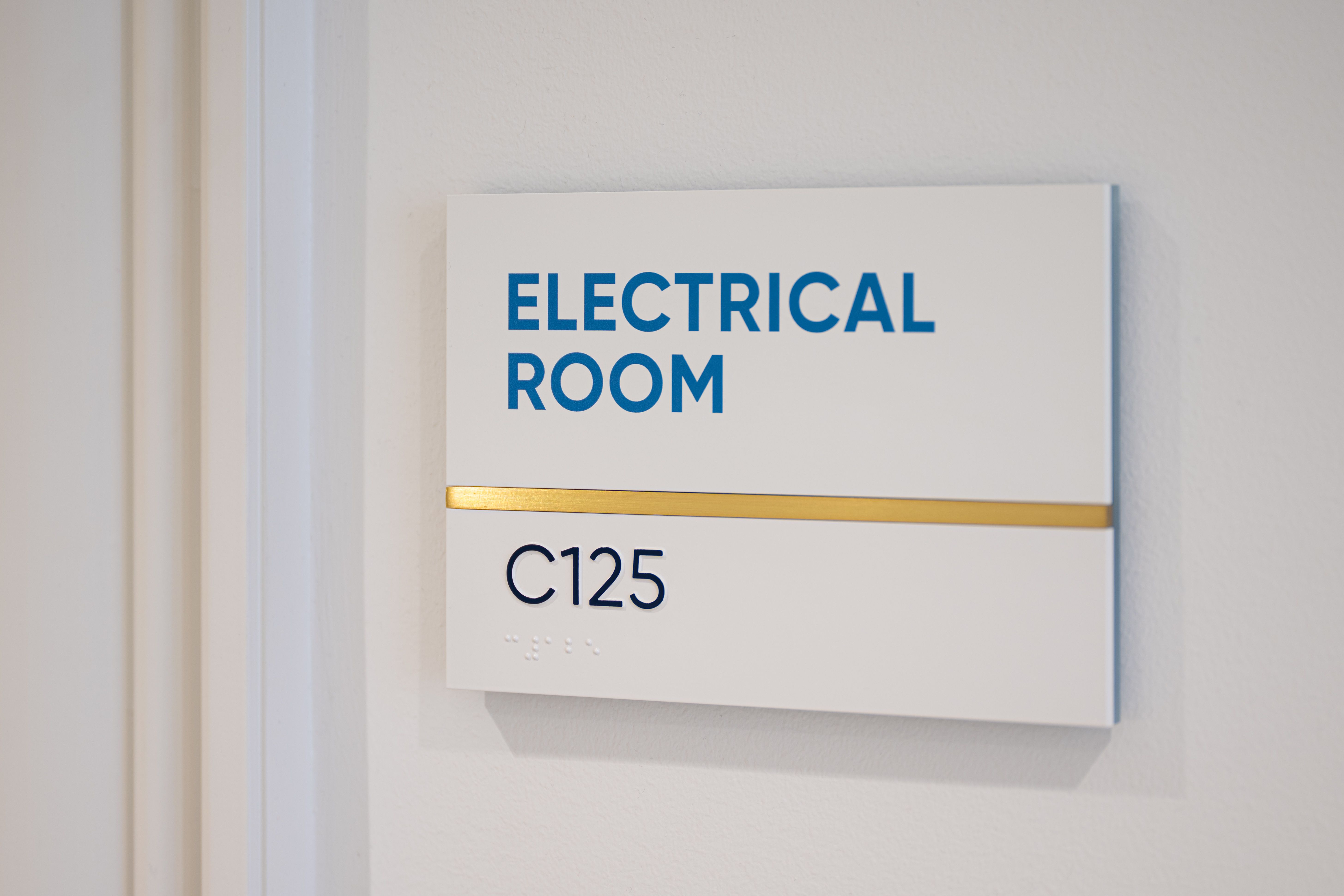 A white tactile room sign with blue edges labeled "Electrical Room" and "C125" with braille, and a brushed brass accent dividing the text.