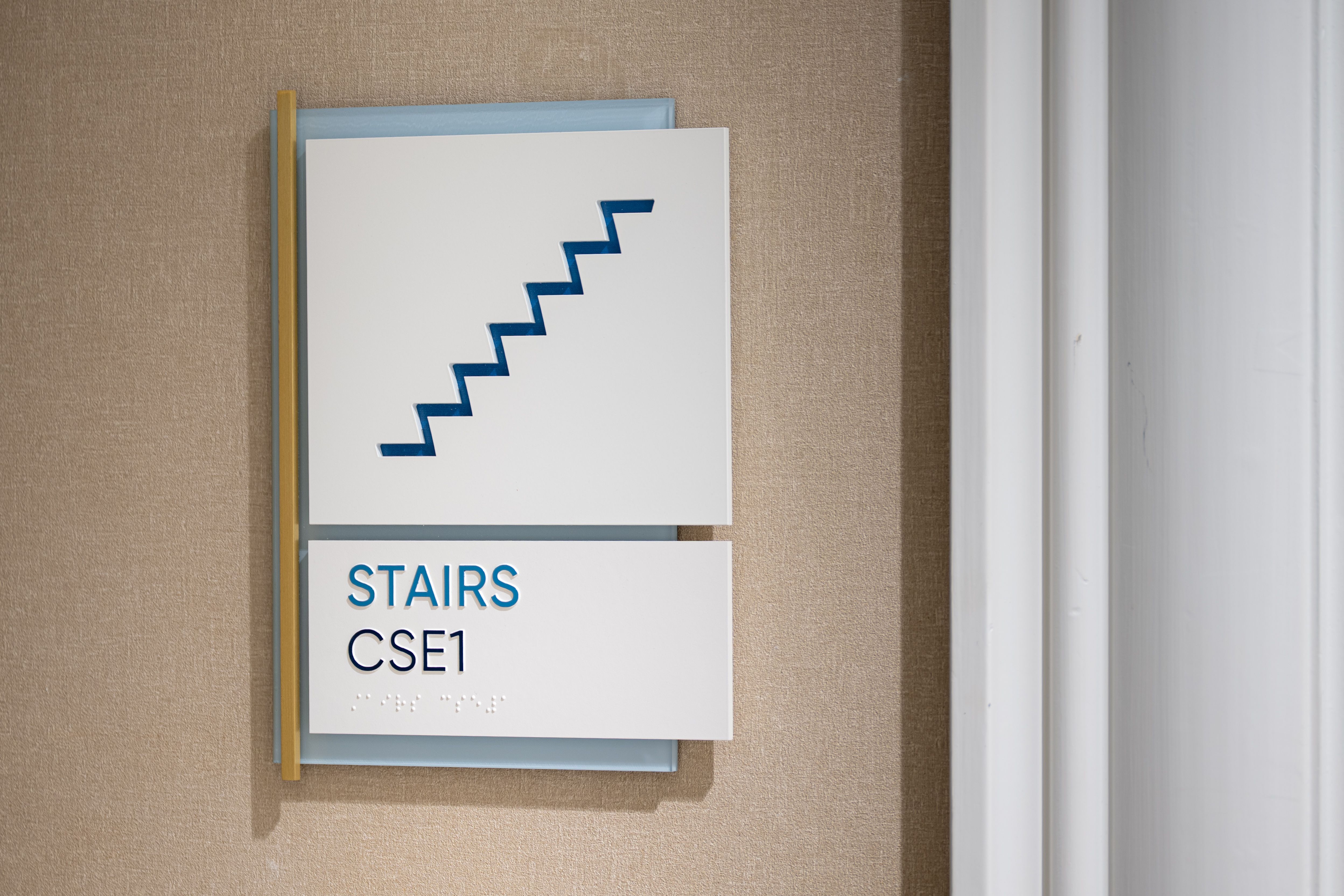 A sign outside a stairwell with raised letters displaying "STAIRS" above the stair number and braille. A stair symbol is laser cut revealing a blue background, while a light blue panel and brushed brass side accent bar add a modern touch.