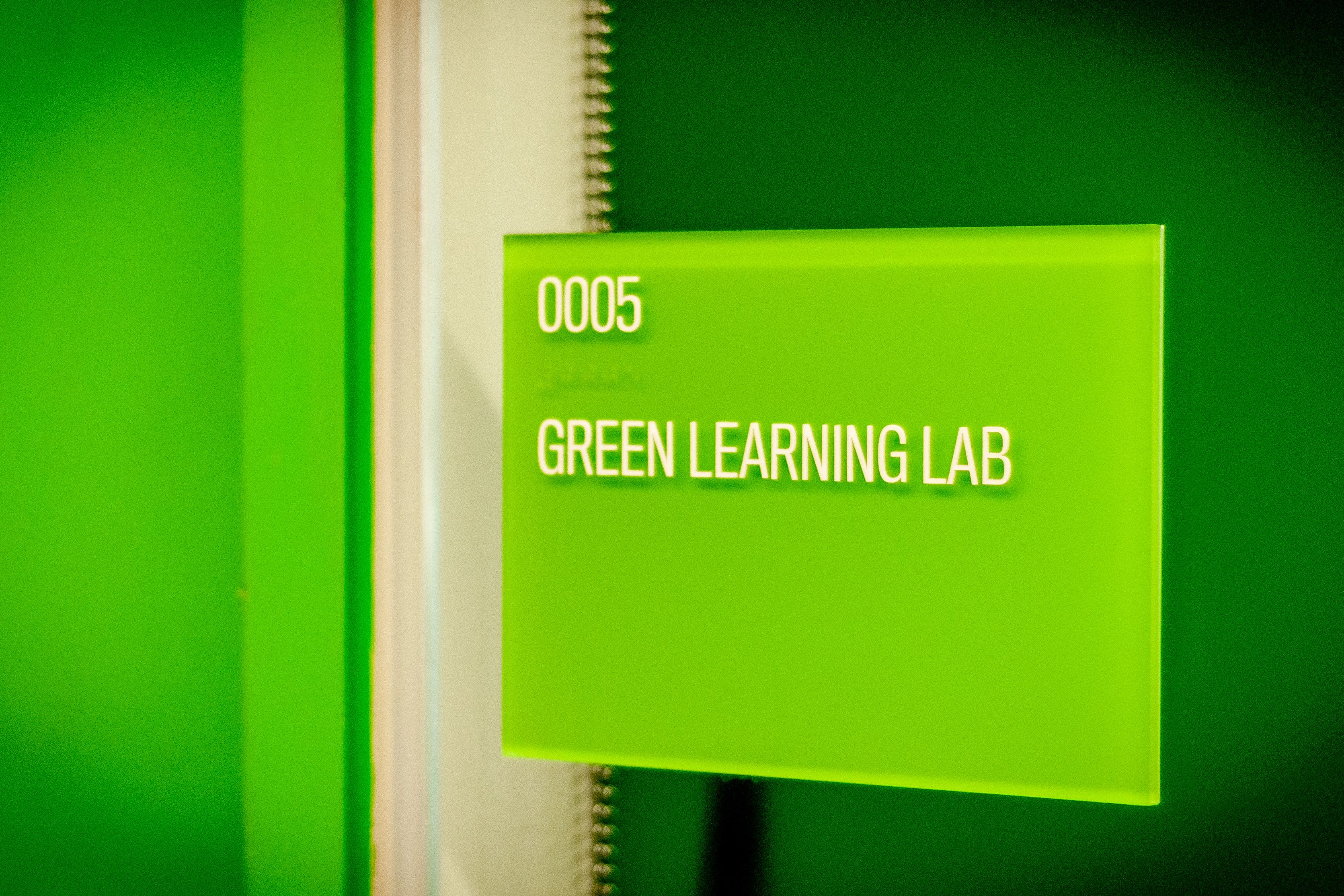 A green acrylic ADA room sign with tactile text and braille is mounted next to a door to the Green Learning Lab.