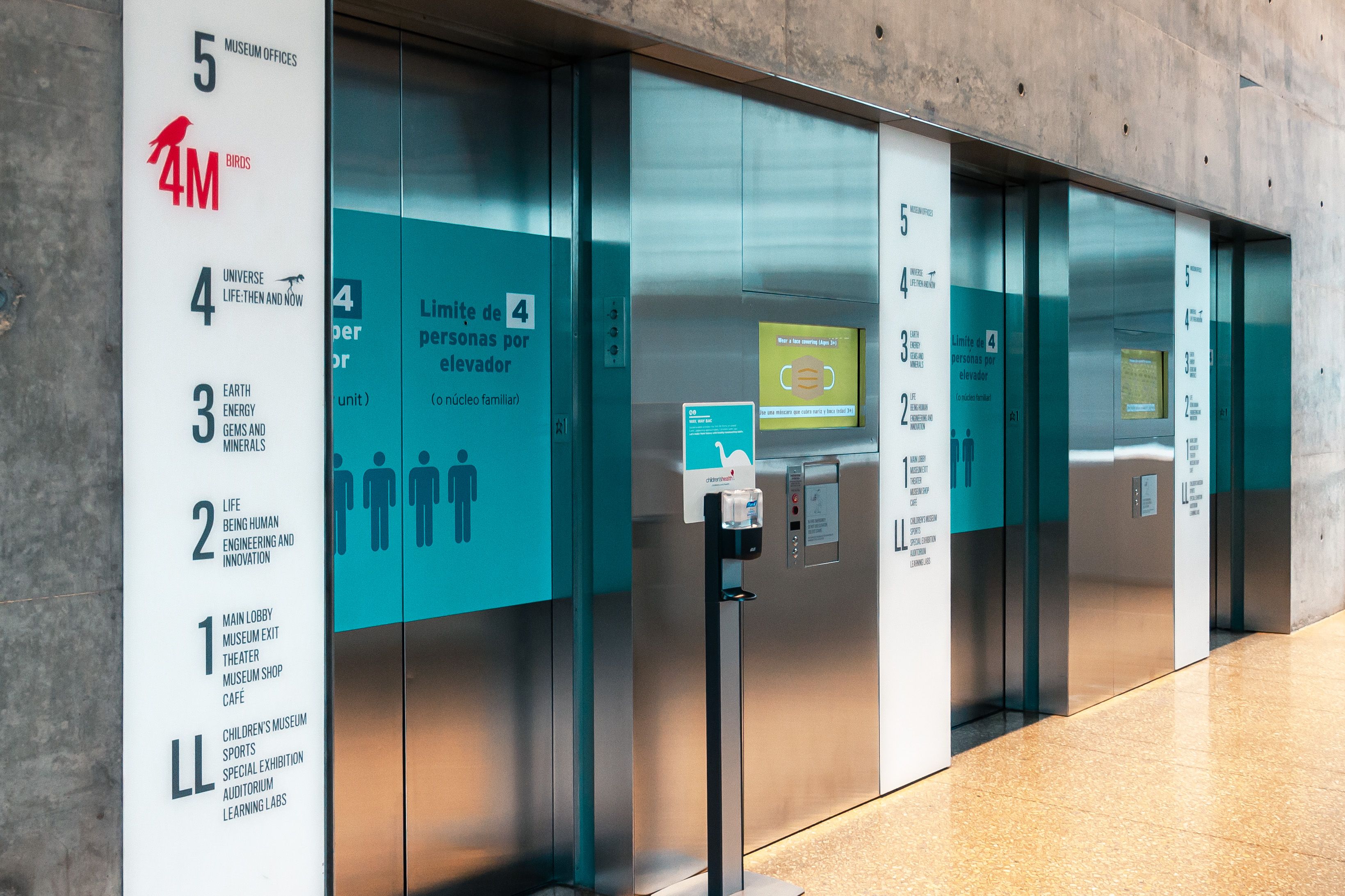 Frosted white acrylic panels mounted next to three elevators show a directory of the Perot Museum's floors and exhibits. The panels have a modern, minimalist design with dark grey text.