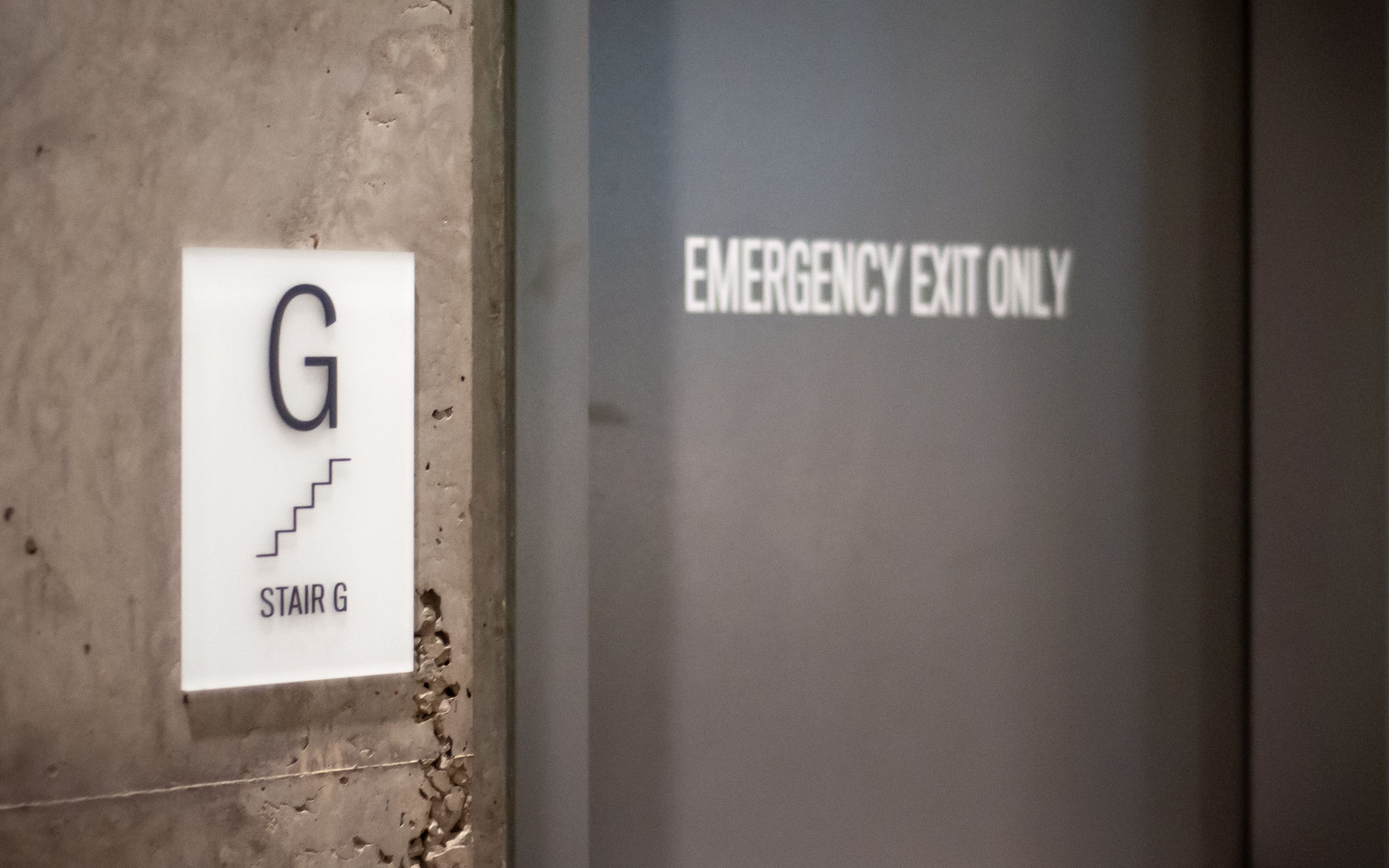 A white and black ADA stair sign with tactile text and braille is mounted next to a stairwell along with a vinyl text on the door saying "EMERGENCY EXIT ONLY"