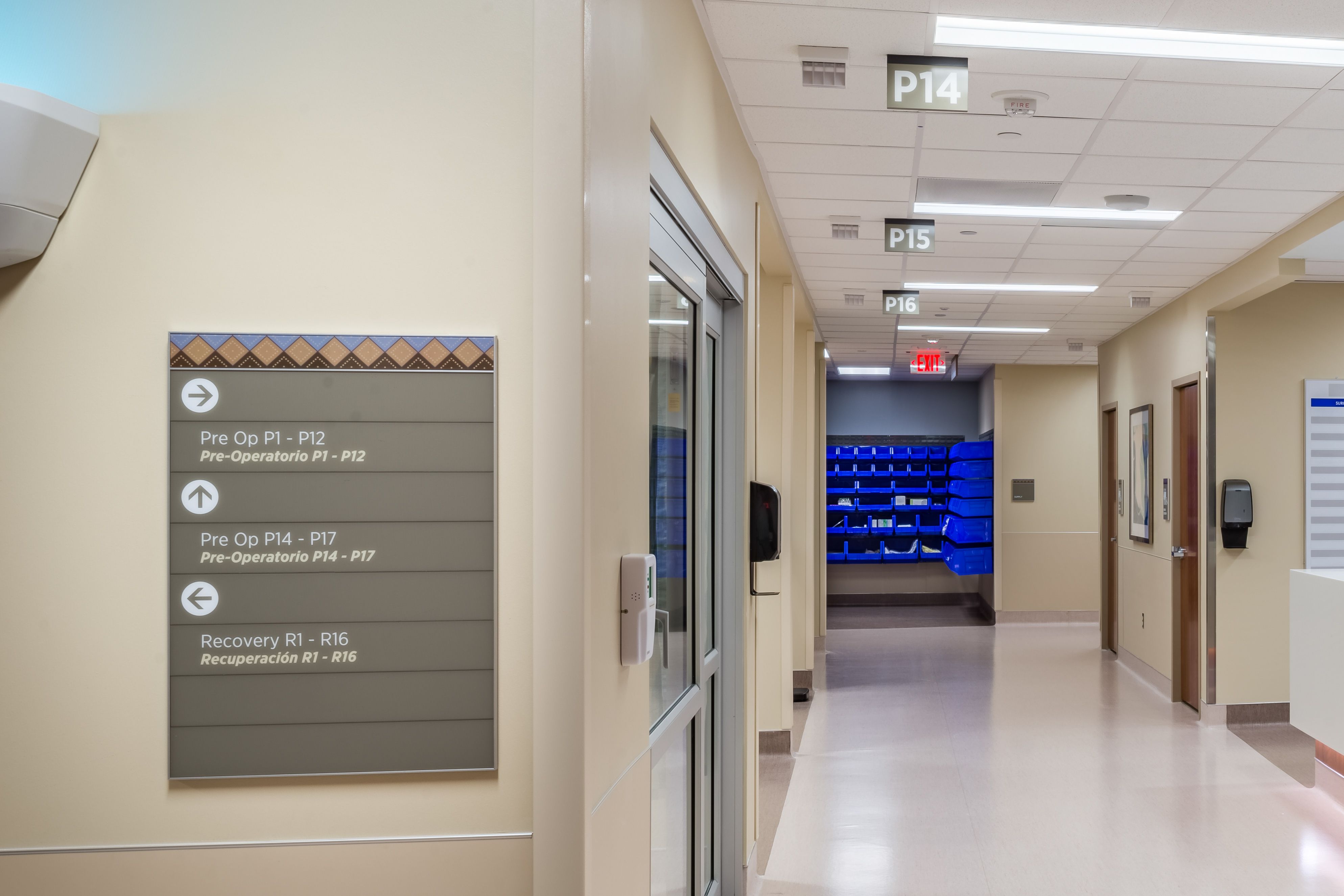 An acrylic wall-mounted directional sign with a patterned header and white text and arrows indicating various departments in a hospital. In the background, flag signs hang from the ceiling, labeling pre-op bays in a hospital.