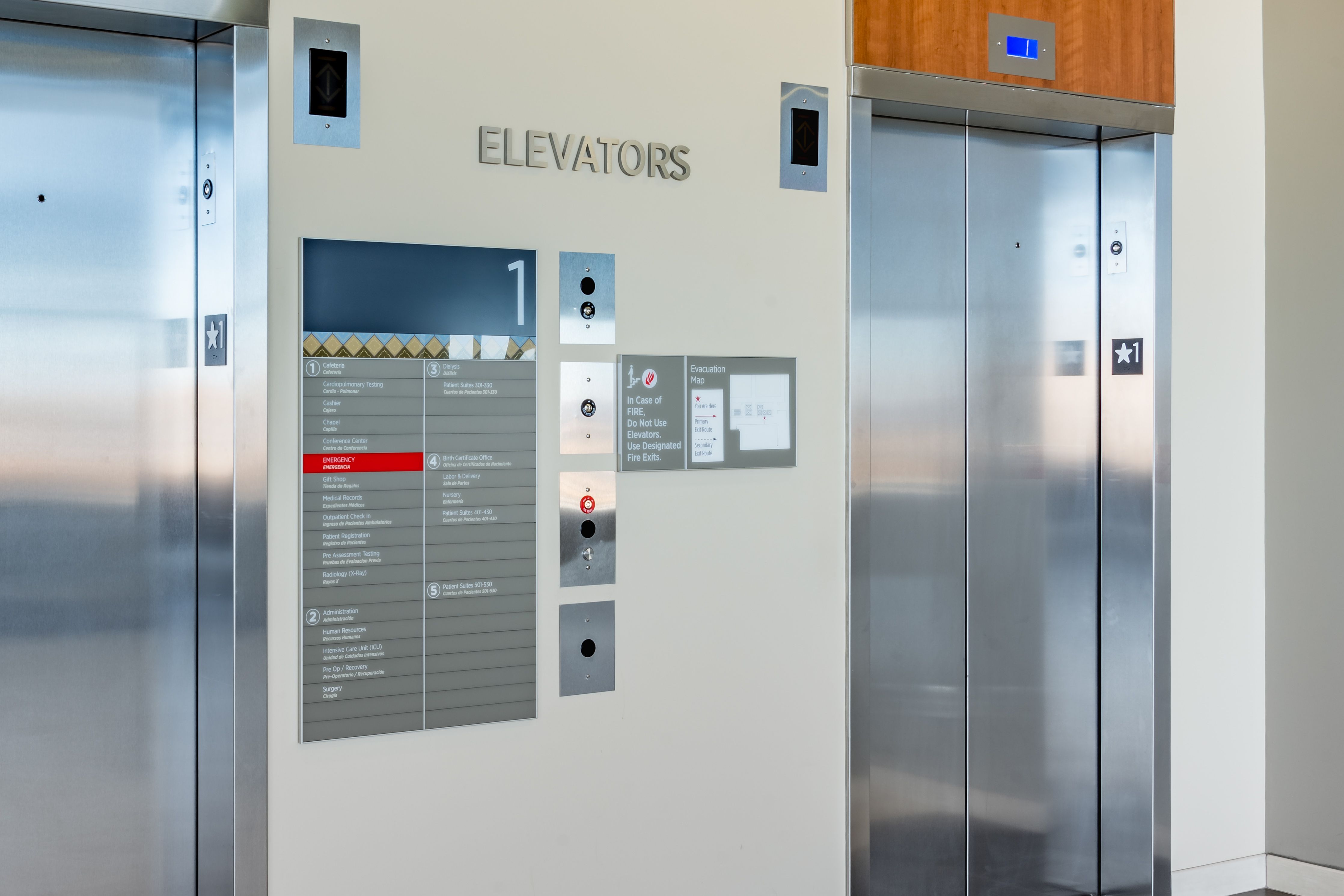 A set of acrylic and aluminum signs, including an elevator evacuation map and a directory listing level destinations, are mounted next to an elevator call button. The large directory sign has a southwest-inspired patterened accent strip and lists departments and their levels.