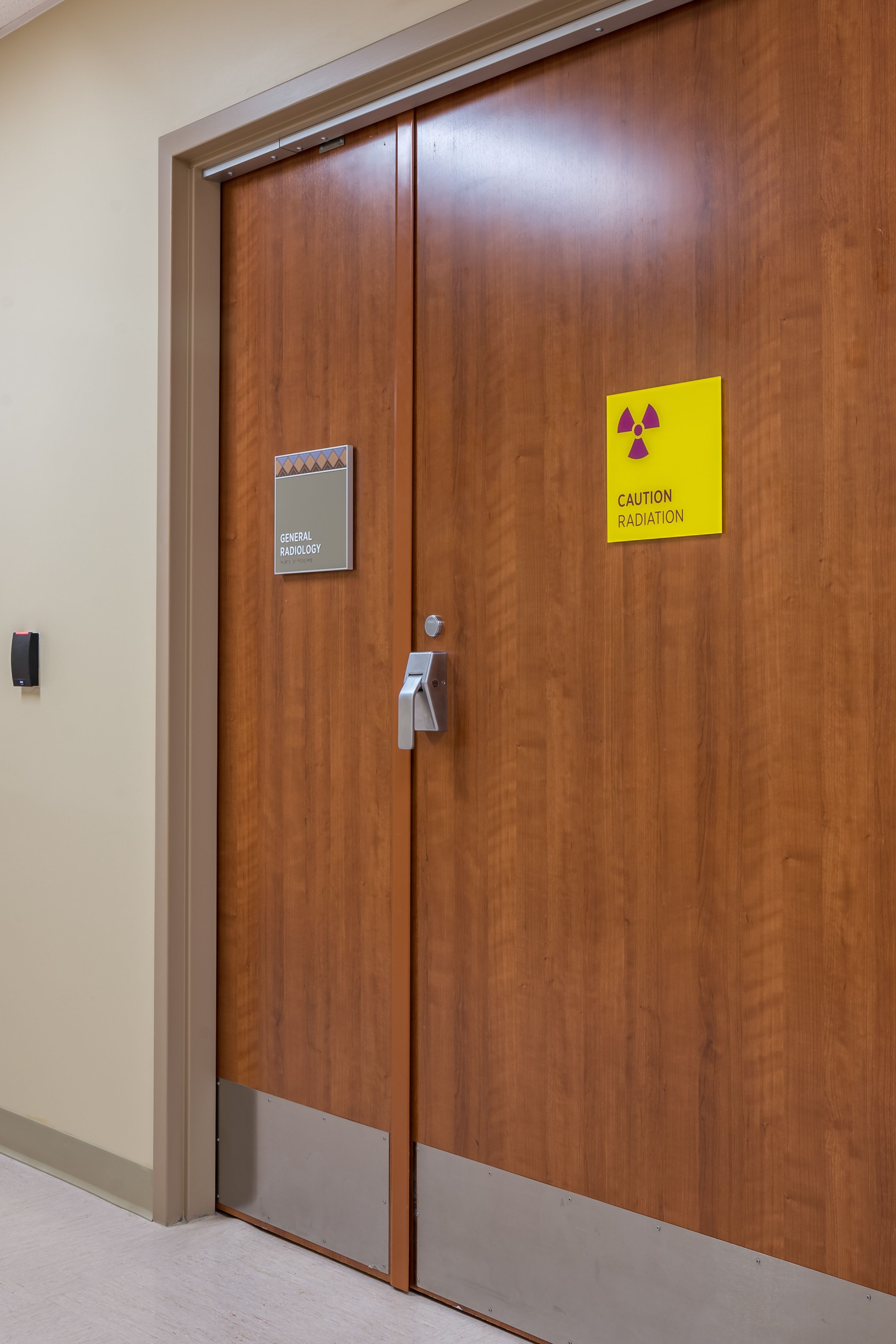 A tactile sign with braille and a decorative patterned header mounted on a door into radiology room, next to a yellow and magenta acrylic "CAUTION RADIATION" sign.