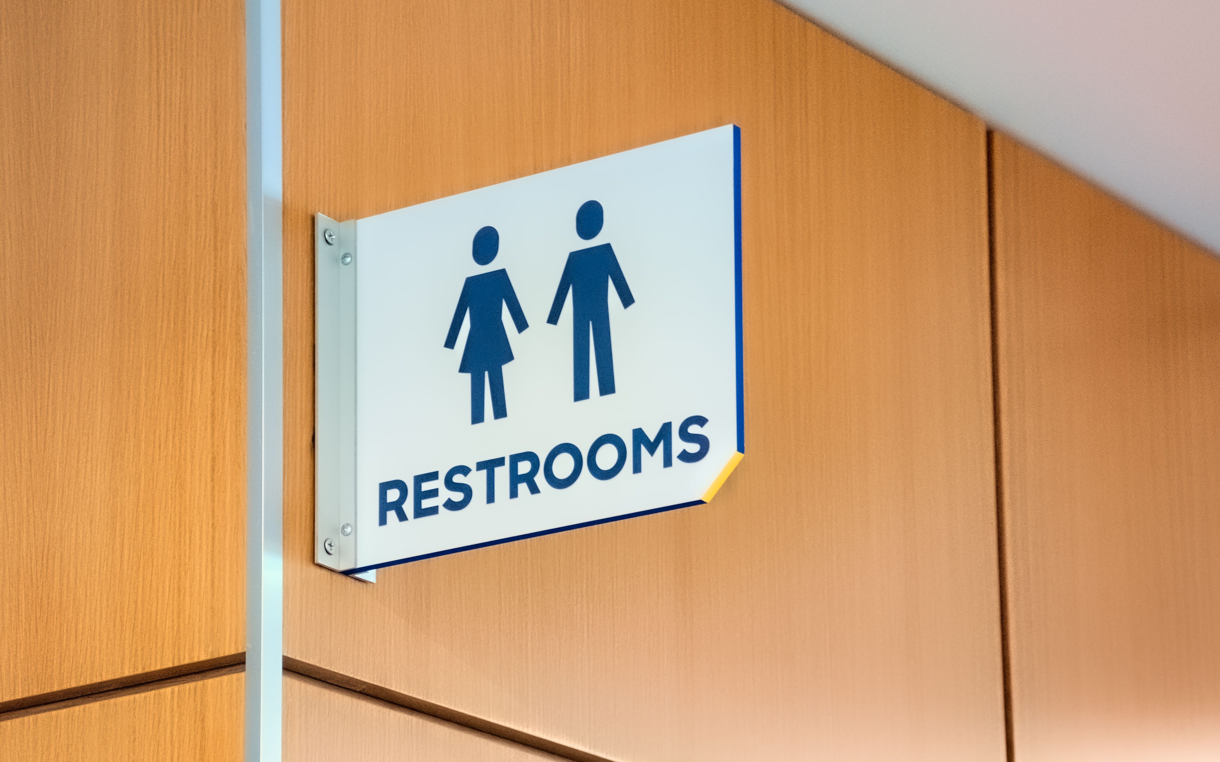 A projecting flag sign with restroom symbols is mounted in a hallway above a restroom.
