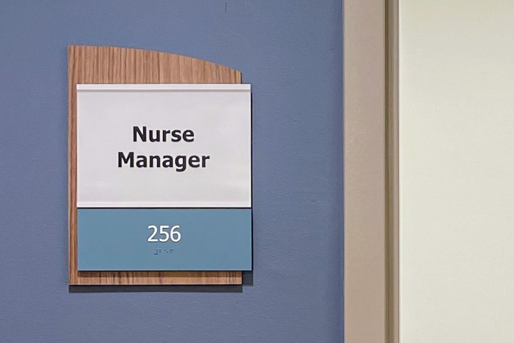 A sign outside a nurse manager's office, displaying the words "Nurse Manager" above the room number "256" and braille, mounted on a blue wall with a wood-accented design.