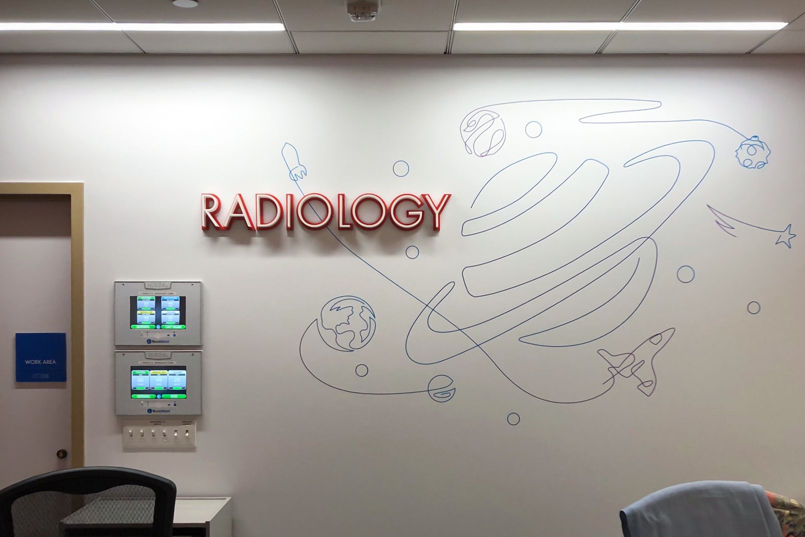 Dimensional laser-cut acrylic letters spell out "RADIOLOGY", mounted on a wall decorated with a whimsical space-themed line drawing. The letters have a layered construction, with a red background and thick white inset face.