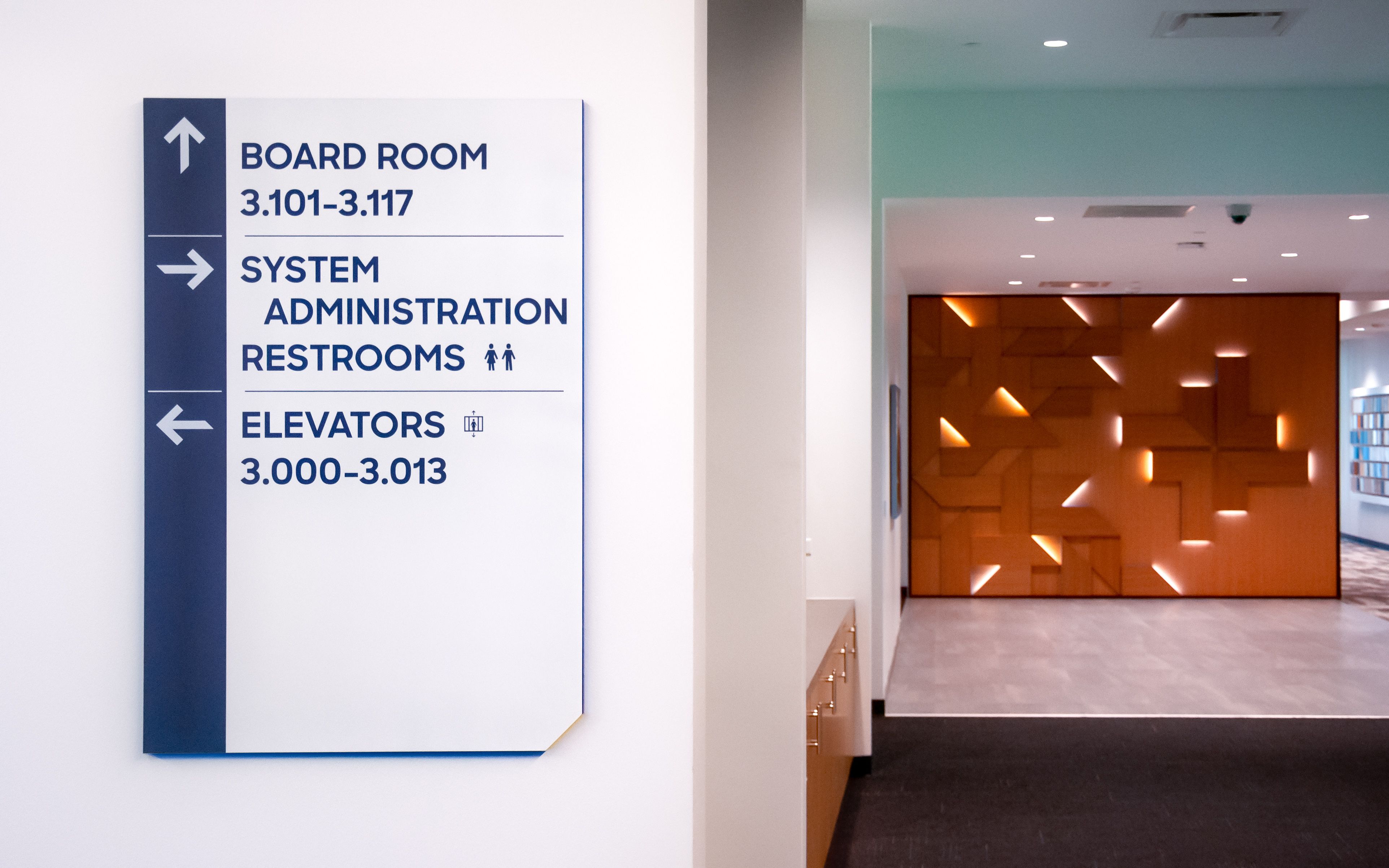 A wall-mounted directional sign with blue and yellow edges points to the various destinations in an administration building.